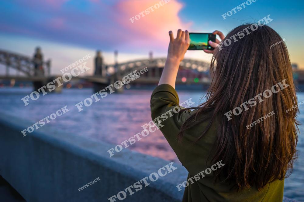 Молодая туристка фотографирует Большеохтинский мост в Санкт-Петербурге на мобильный телефон во время прогулки по набережной. Величественная архитектура моста, огни города и атмосфера северной столицы России создают живописный городской пейзаж. Фотография передаёт настроение путешествия, современный городской туризм и красоту знаменитых мостов Петербурга. Изображение идеально подходит для публикаций о путешествиях по России, туризме в Санкт-Петербурге, городских прогулках, архитектуре мостов и туристических достопримечательностях.